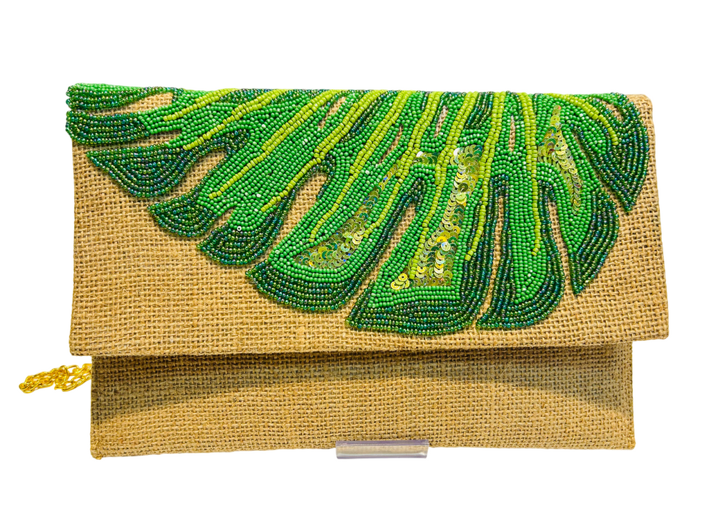 The Hand-Embroidered Monstera Jute Evening Bag is a rectangular tan eco-friendly clutch with textured fabric, green beadwork and sequins in a leafy design on the flap, plus a small gold chain attached to the side.