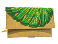 The Hand-Embroidered Monstera Jute Evening Bag is a rectangular tan eco-friendly clutch with textured fabric, green beadwork and sequins in a leafy design on the flap, plus a small gold chain attached to the side.