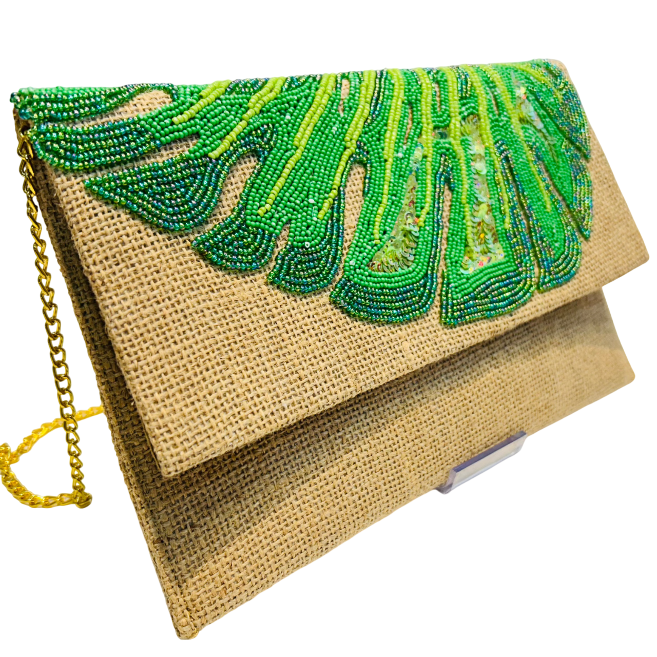 The Hand-Embroidered Monstera Jute Evening Bag is a tan, eco-friendly purse with a gold chain strap, featuring intricate green and yellow beaded leaf designs—a perfect tropical accessory for any occasion.